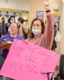 woman with sign saying "Low pay makes PCA Shortage" indoors