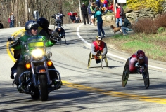 several wheelchair racers going downhill and police motorcycle