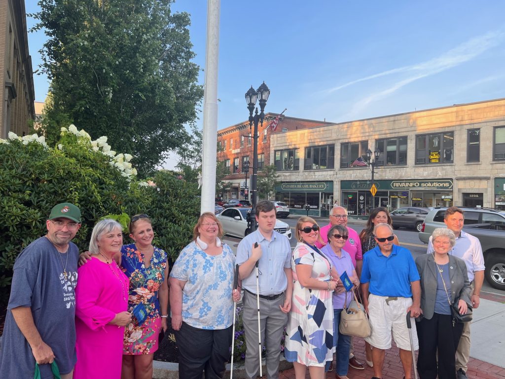 The Disability Commission, Mayor and State Reps pose in front of flag pole. 