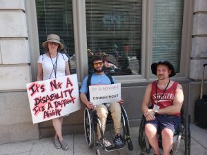 Woman standing with sign: Not a disability, a different Ability. Man in wheelchair with sign Indivisible Connecticut. Second man in photo in wheelchair with no sign.